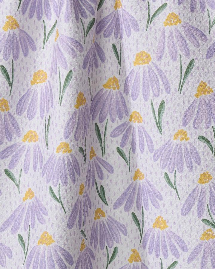 A close-up of Hufton Studio's Upside Down Daisy tea towel, made from recycled materials, shows soft fabric folds and a white background decorated with a repeating pattern of purple daisies with yellow centers and green leaves.