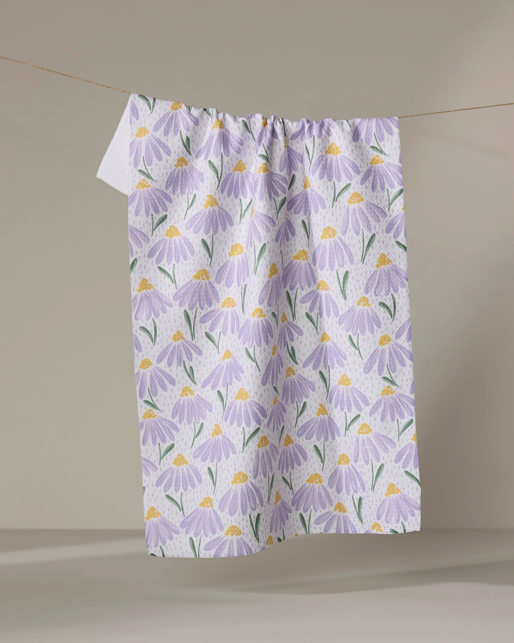 The Upside Down Daisy tea towel by Hufton Studio, featuring purple flowers and green leaves, is made from recycled materials and hangs on a clothesline against a neutral background.