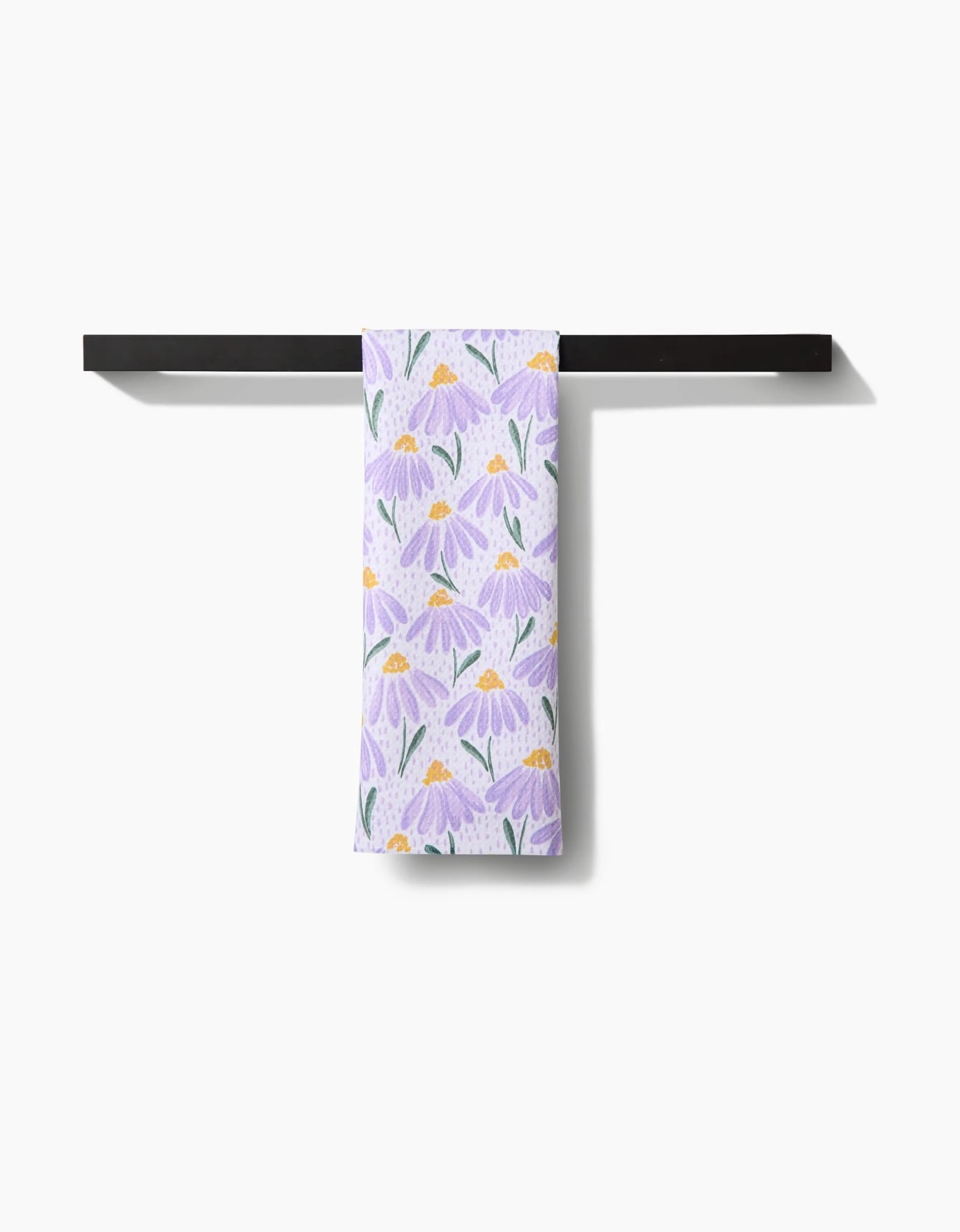 The Upside Down Daisy hand towel by Hufton Studio, featuring a purple daisy pattern and made from recycled materials, hangs neatly on a simple black horizontal towel bar against a plain white background.