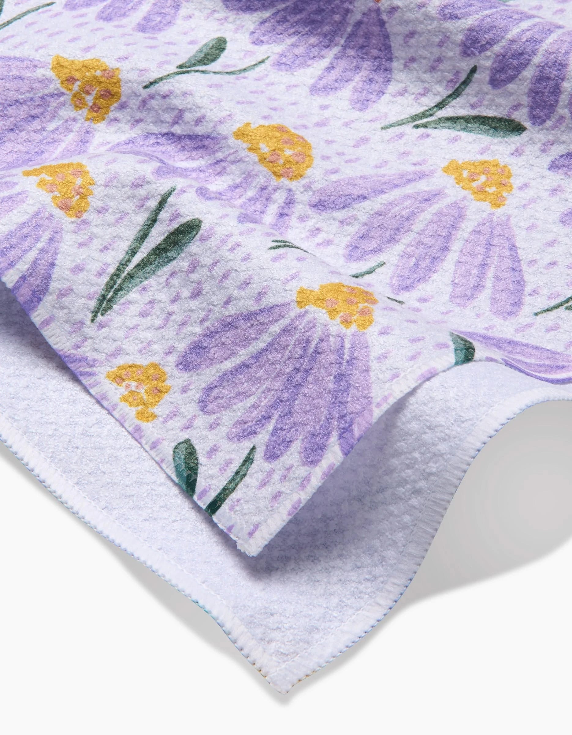 A close-up of the Upside Down Daisy Tea Towel by Hufton Studio shows a floral pattern with purple petals, yellow centers, and green leaves on a light background. Quality Geoweave fabric is highlighted at the edge and textured underside.