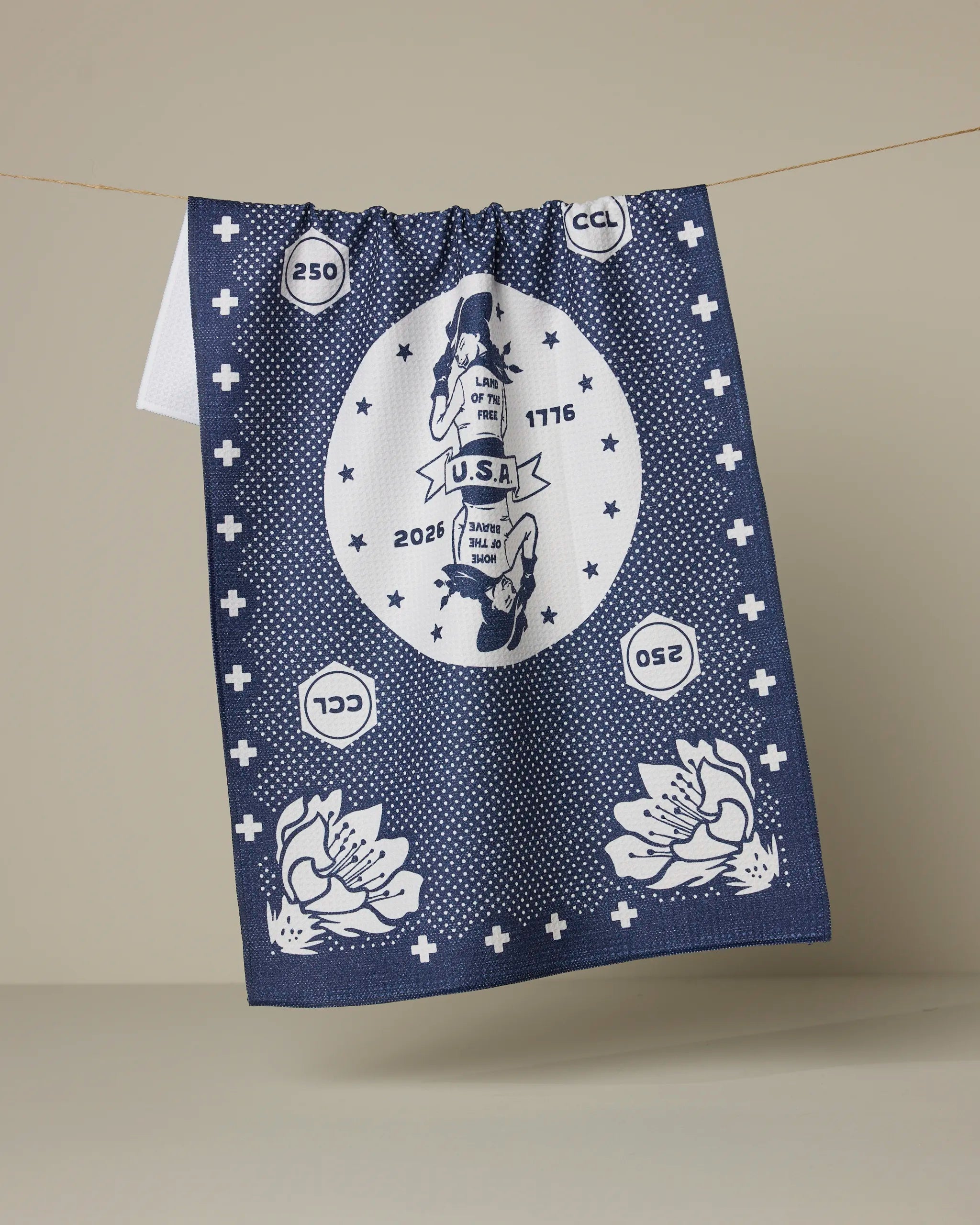 A navy blue and white ultra-absorbent Geometry USA Cowgirl towel hangs on a line, featuring a central circle with "U.S.A." and "2026," numbers, "CCL," and floral accents at the corners.