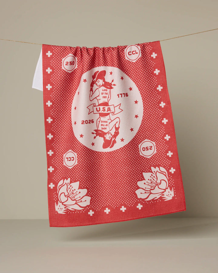 A red and white Geometry "USA Cowgirl" kitchen tea towel hangs on a line, ultra-absorbent and featuring vintage-style graphics—floral designs, stars, numbers, and "U.S.A." with two hands and ribbon illustrations.