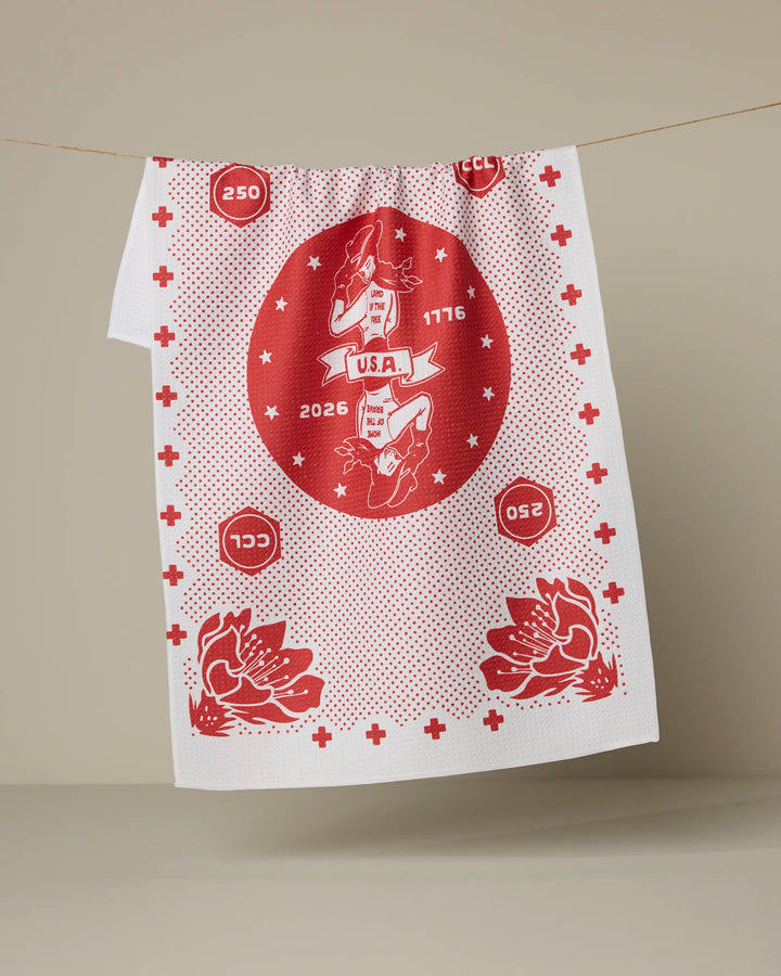 The Geometry USA Cowgirl kitchen tea towel, made from recycled materials, is white with red graphics featuring a central Texas map, the years 1776 and 2026, circled numbers, and two floral designs at the bottom.