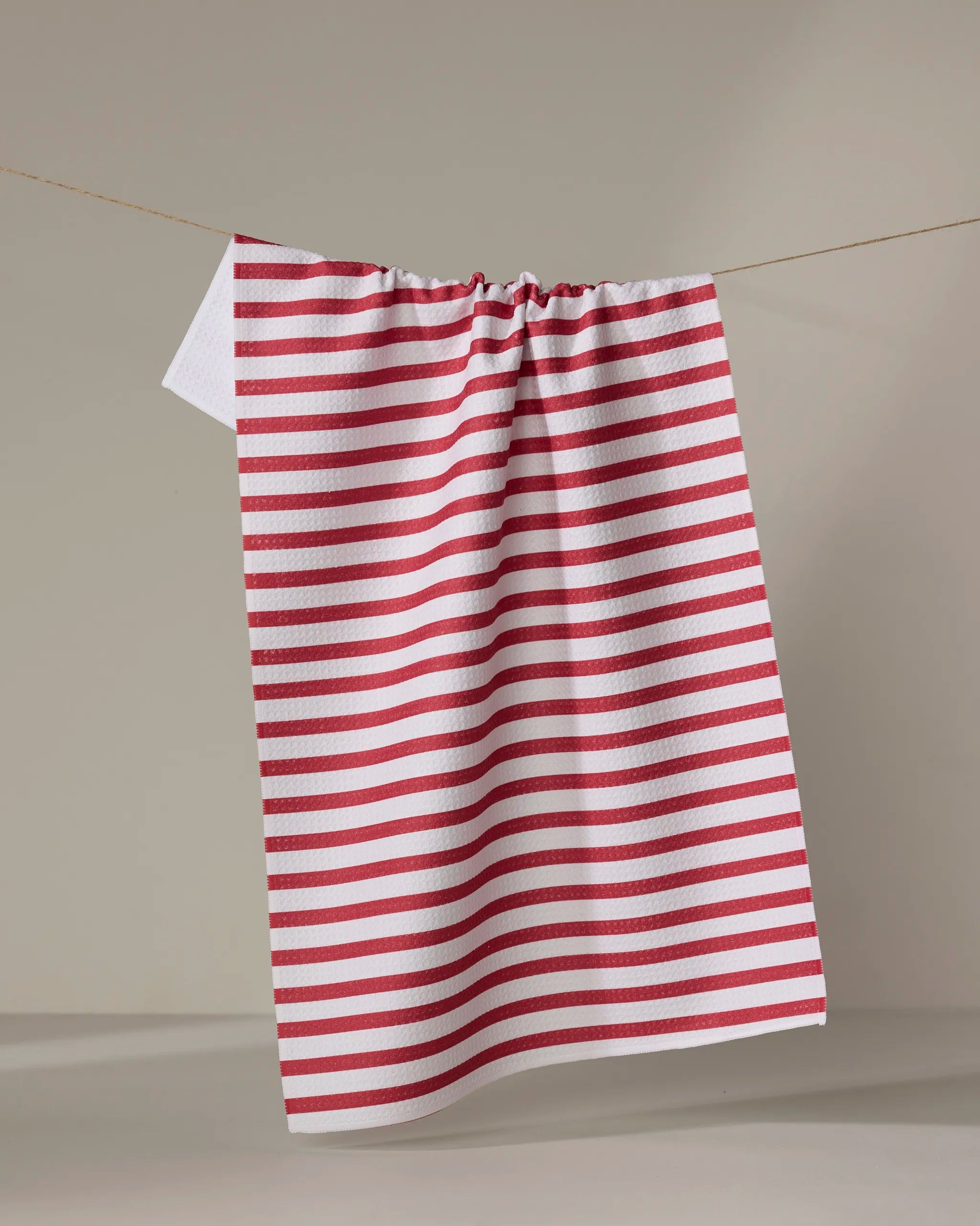 The Mindfulnice USA Stripes quick-drying towel, featuring red and white stripes, hangs on a clothesline against a beige background.