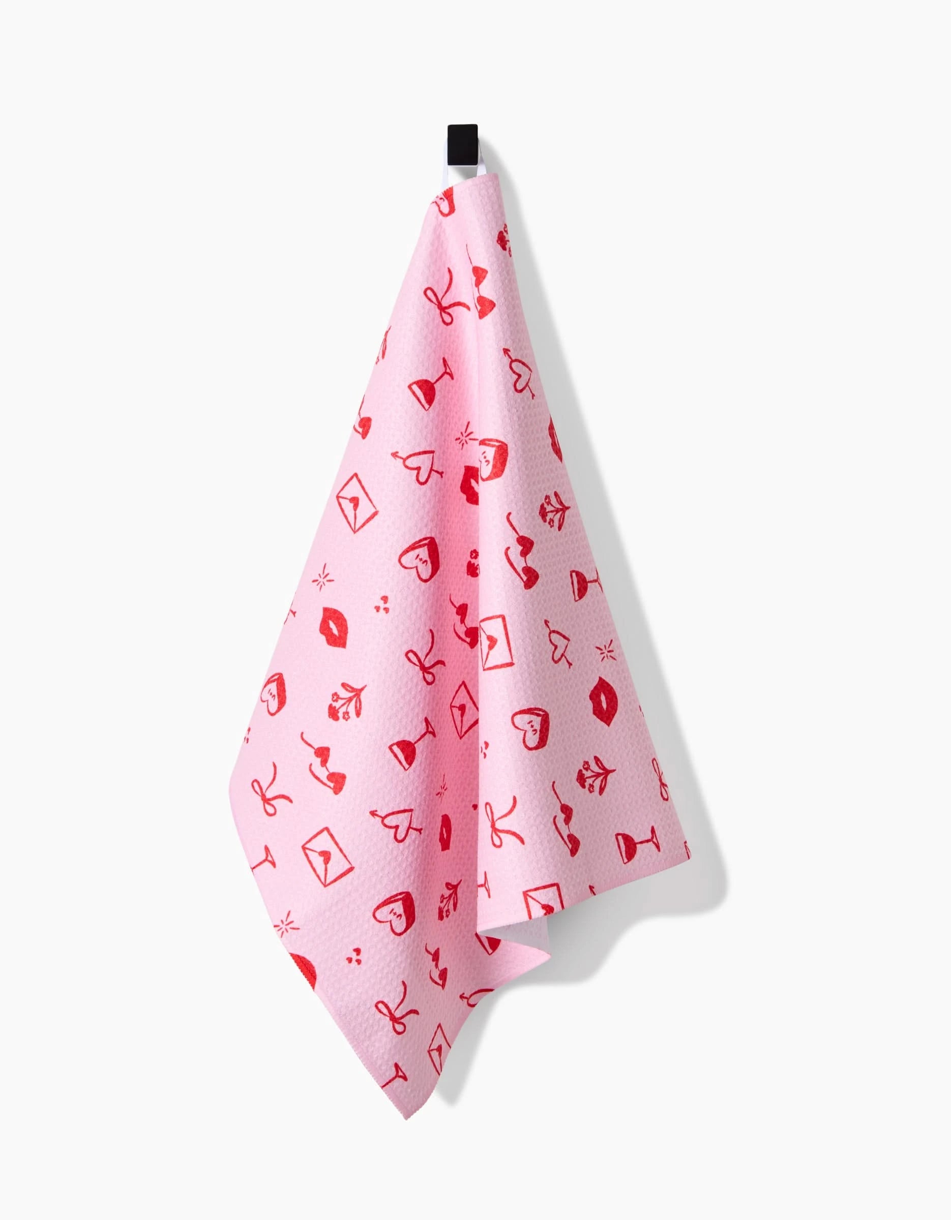 The V Day Doodles tea towel by Laura Supnik, featuring red lips, hearts, envelopes, and drinks on ultra-absorbent pink fabric, hangs from a black hook against a white background.