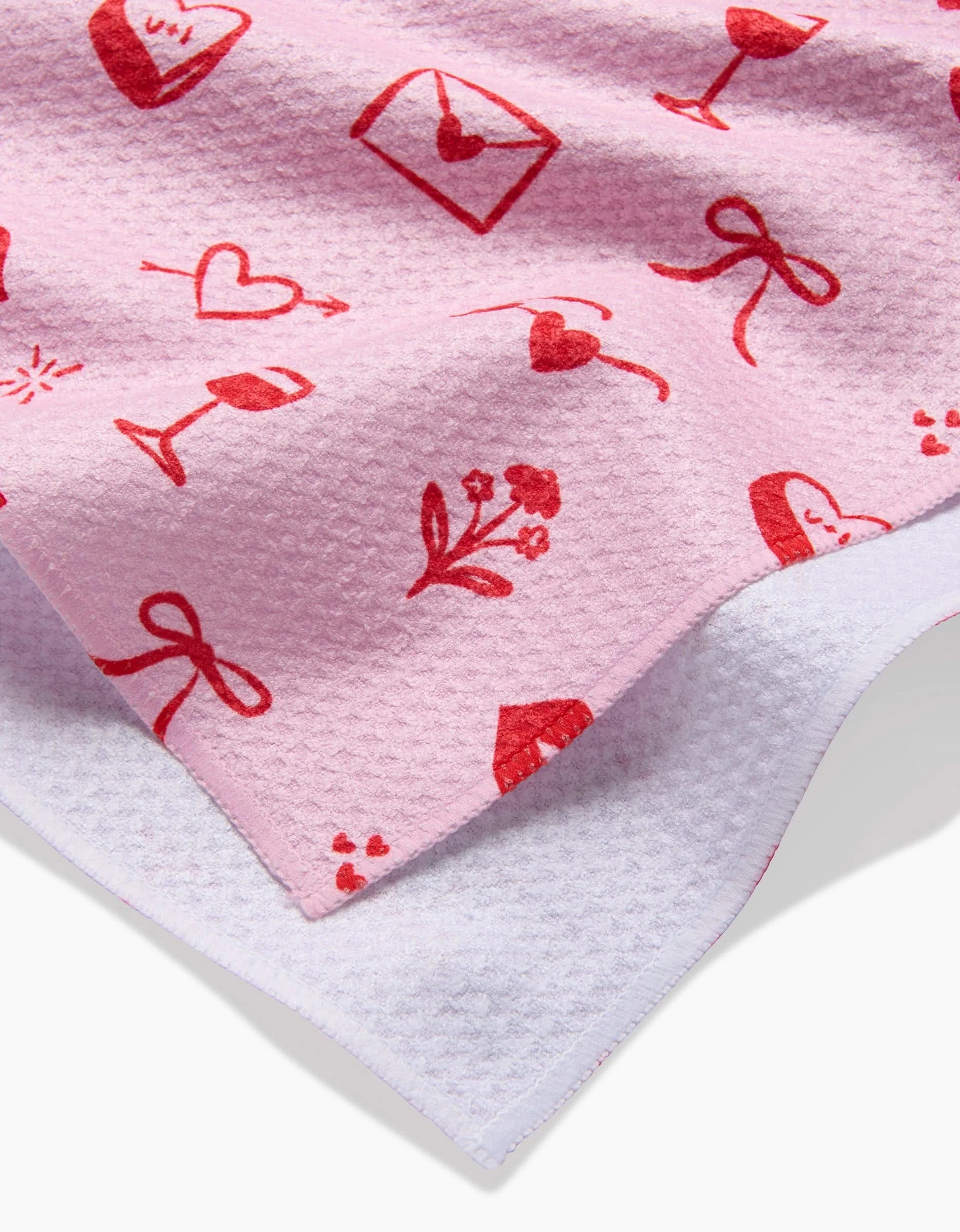 A close-up of Laura Supniks V Day Doodles tea towel—pink, textured, and made from recycled materials—features red Valentines illustrations like hearts, bows, wine glasses, flowers, and love letters on white fabric.