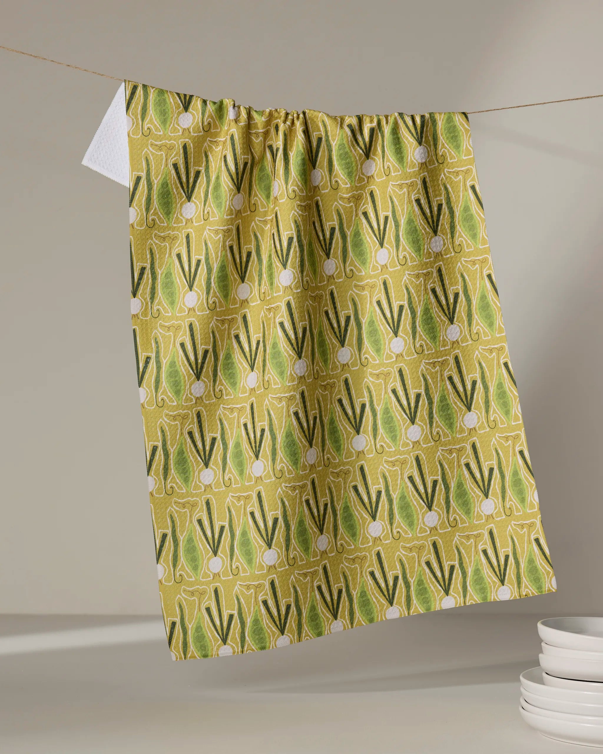 The "Veg on Parade" tea towel by Lizzie Clark, featuring a yellow background and green onion pattern, hangs on a line above a white surface with stacked white dishes in the corner.