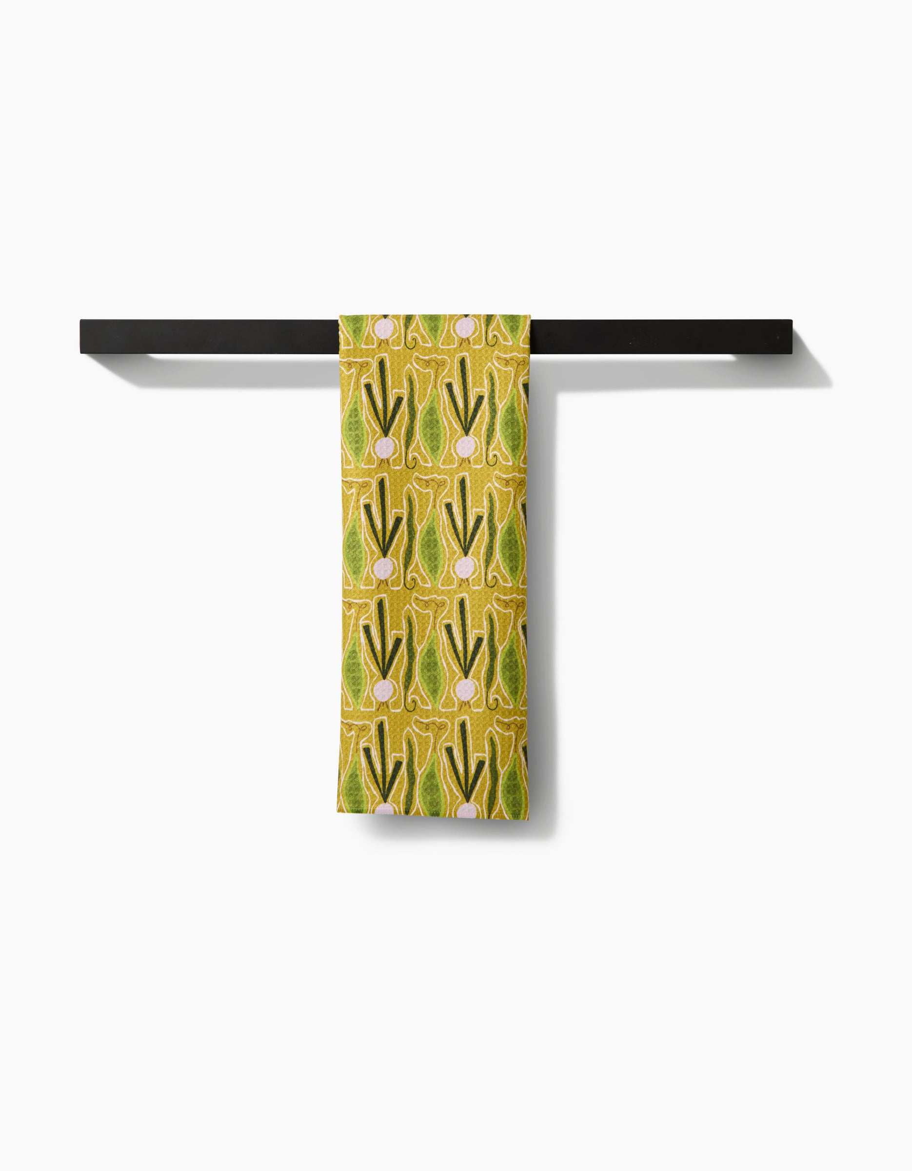 The Veg on Parade tea towel by Lizzie Clark, featuring a retro yellow and green vegetable pattern and made from recycled materials, hangs over a black bar against a white background.