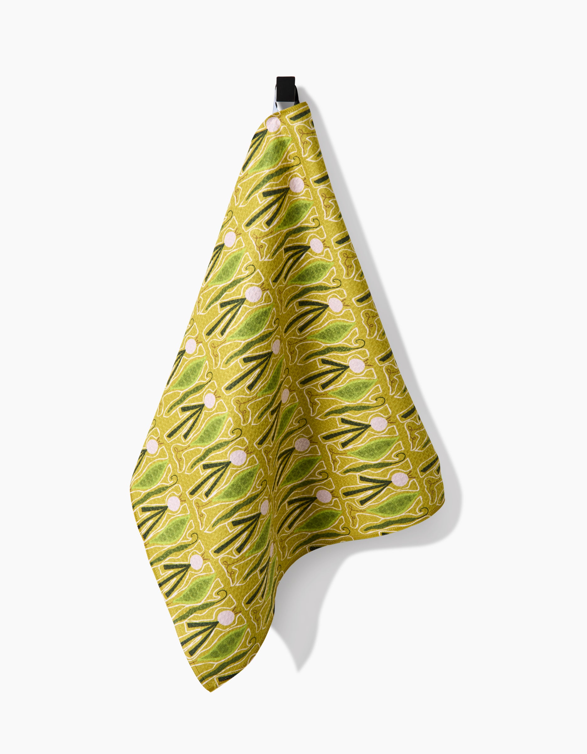 A green and yellow patterned Veg on Parade tea towel by Lizzie Clark, made with Geoweave fabric and pink and white circular accents, hangs from a black hook against a white background.