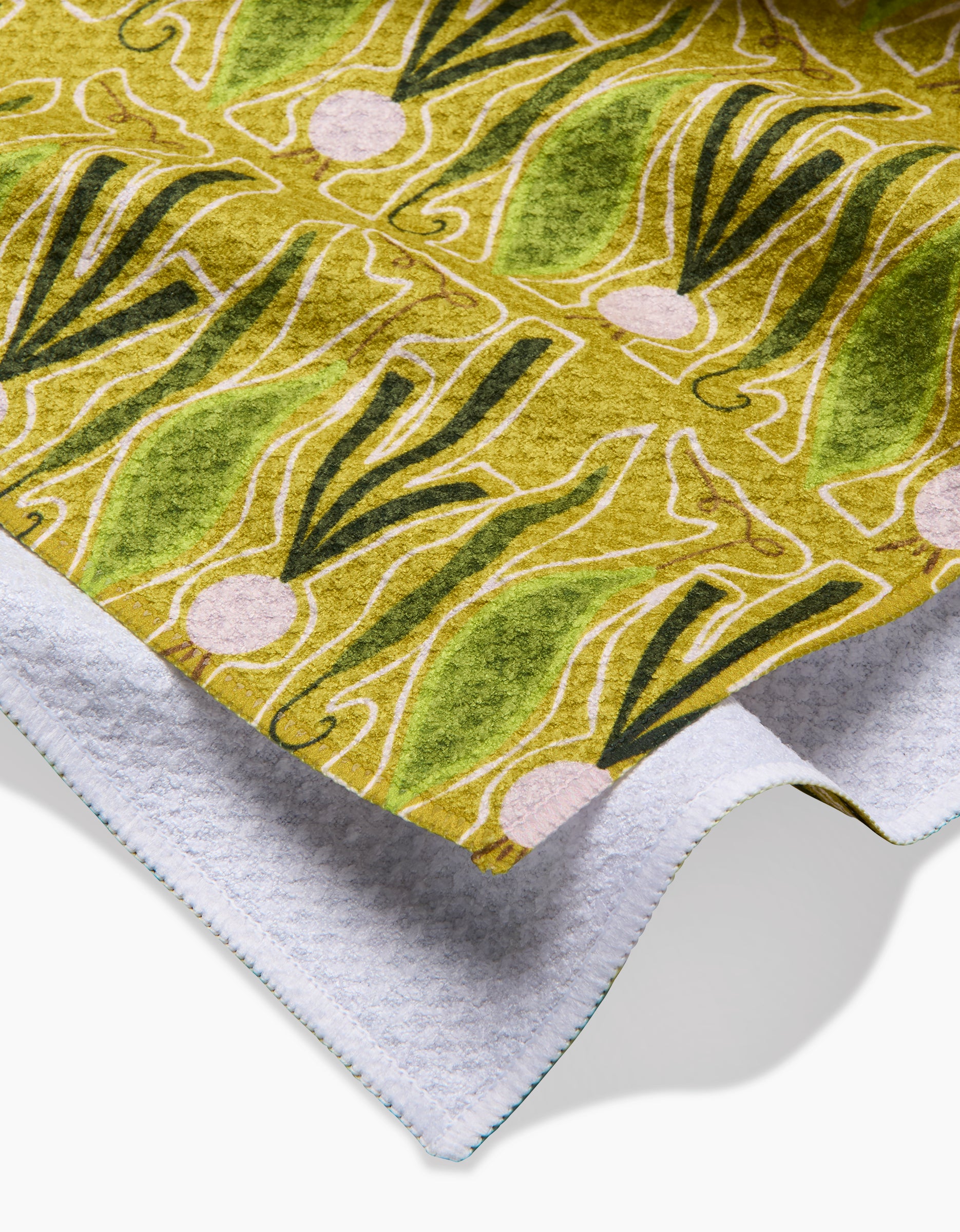 A close-up of the Lizzie Clark Veg on Parade tea towel, showcasing its green and yellow geometric pattern with abstract scallion shapes, made from Geoweave fabric and a partially visible white textured underside.