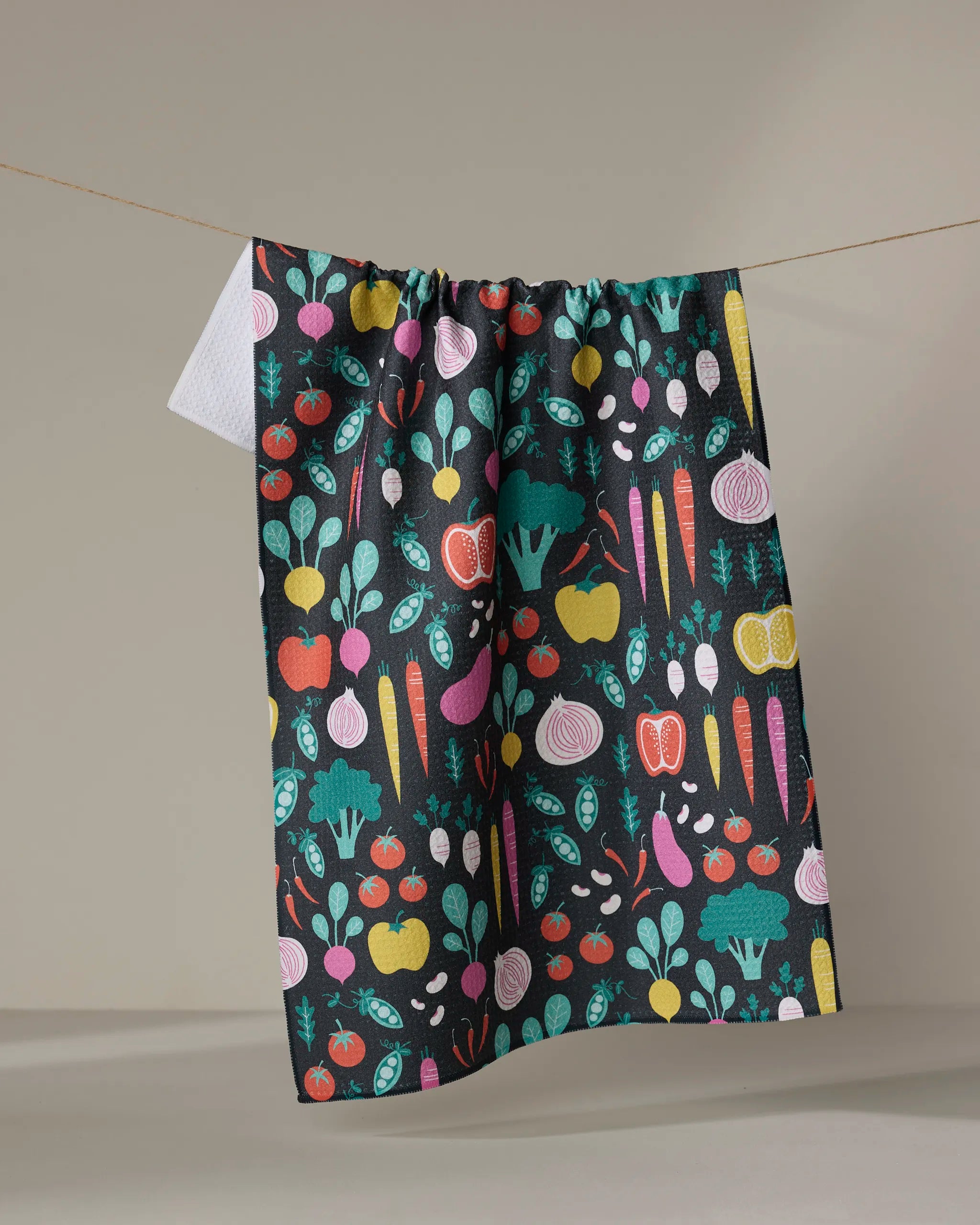 The "Veggies" kitchen towel by Megan Kampa, featuring a vibrant pattern of carrots, tomatoes, apples, broccoli, turnips, and peas, hangs against a beige background.