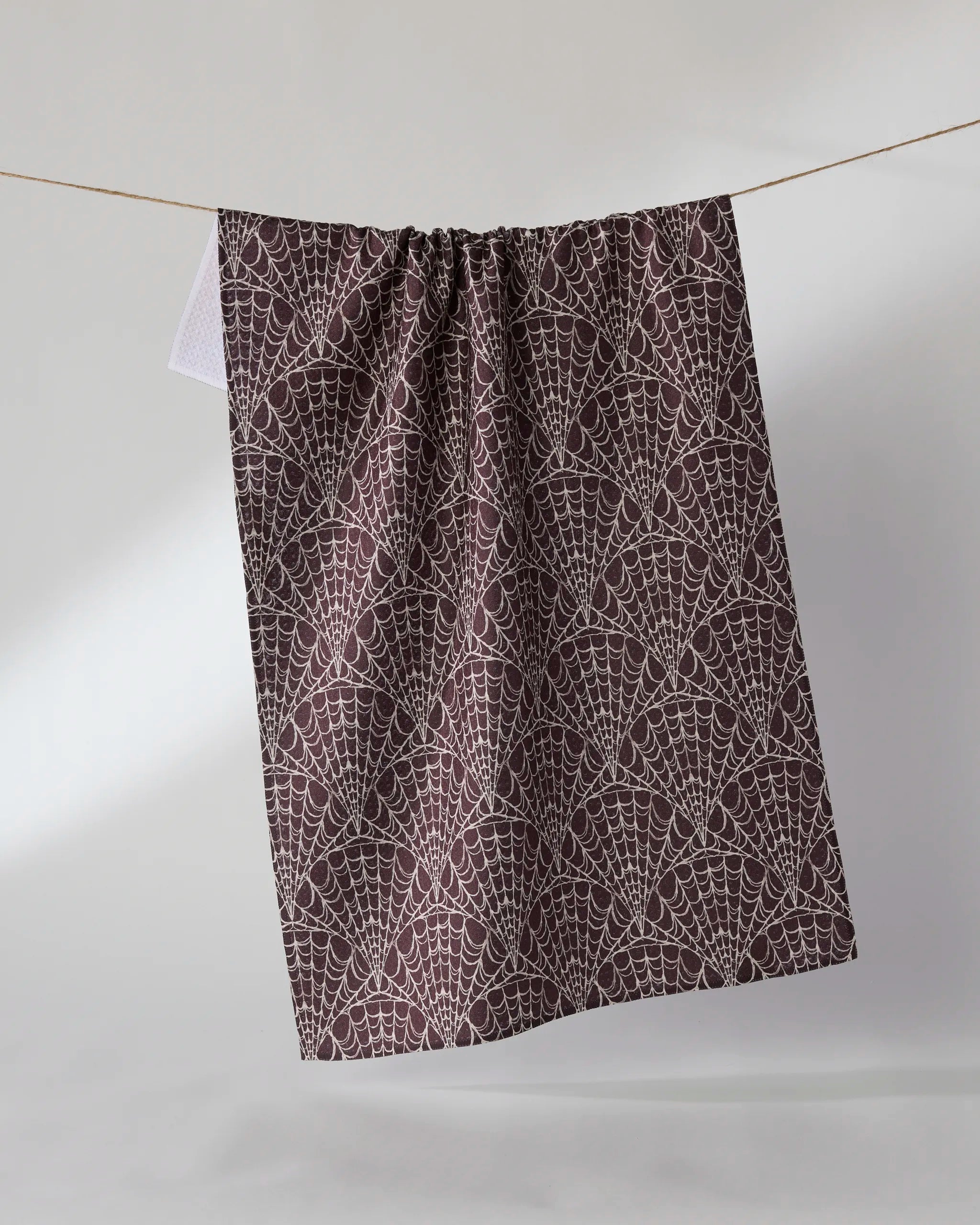 The Geometry “Velvet Cobwebs” tea towel, made from recycled materials in dark brown with a white cobweb pattern, hangs from a string against a light background.