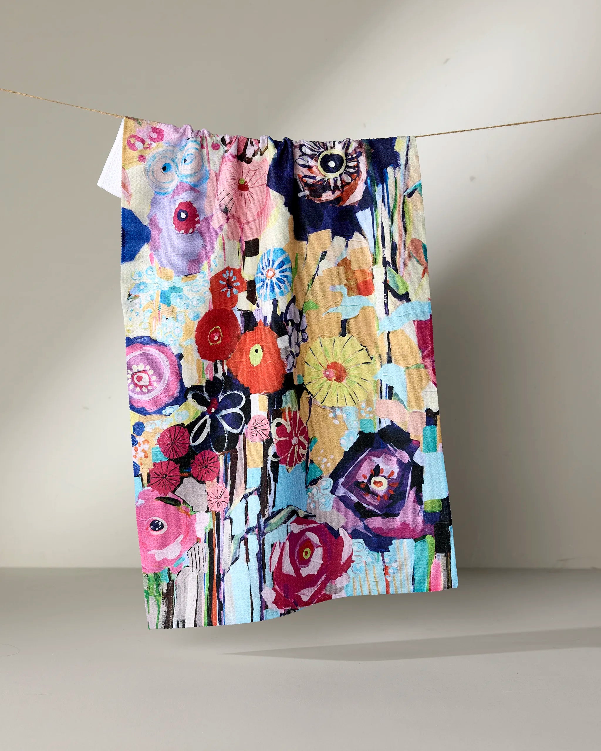 Vera, a vibrant kitchen towel by Ania Zwara Art, features abstract floral patterns and is crafted from recycled materials. It hangs on a line against a neutral background.