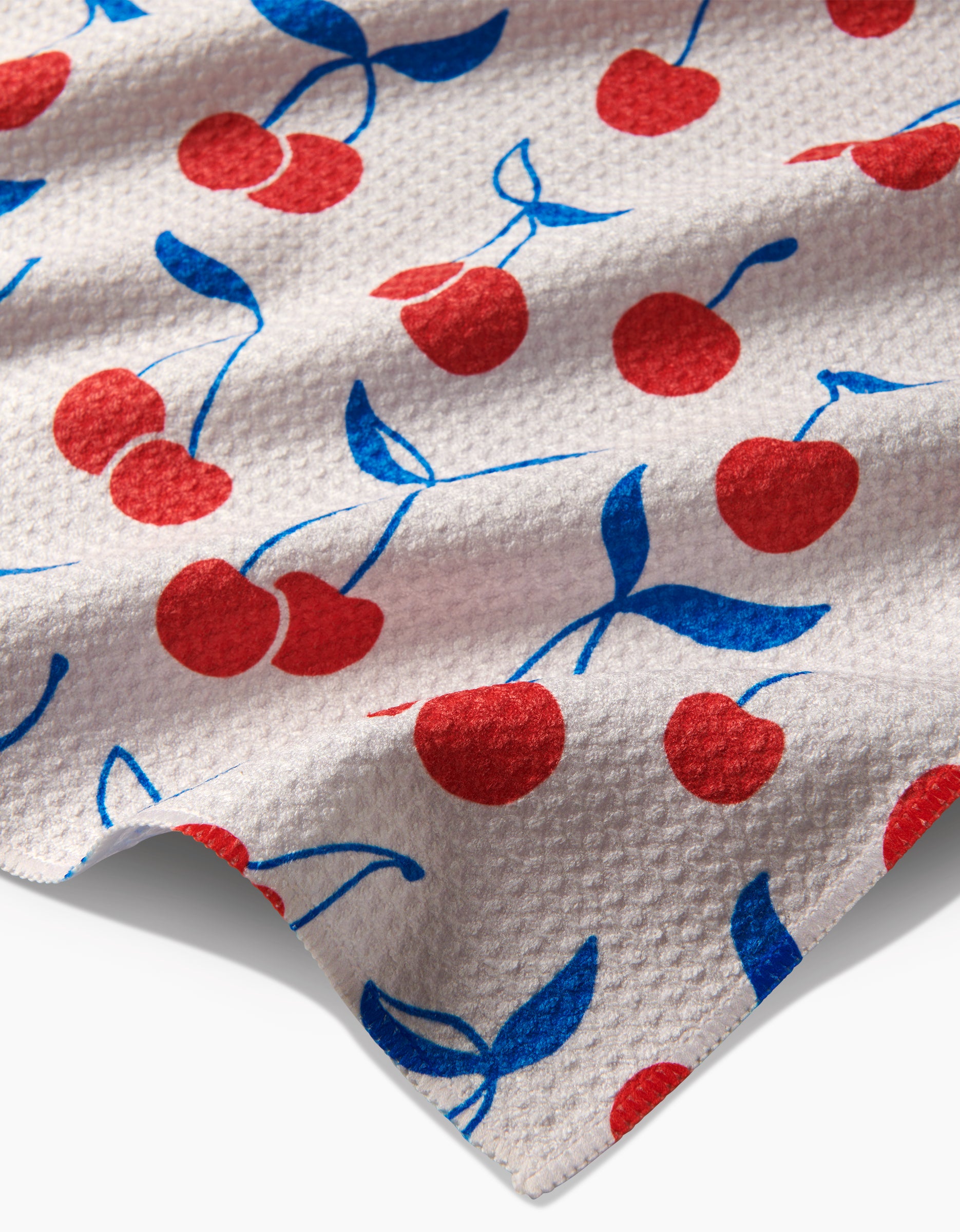 The Kayla Ann Very Cherry tea towel features textured white Geoweave fabric with a red cherry pattern and blue stems and leaves, shown close up with a slightly wrinkled finish.