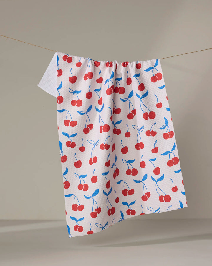 The Kayla Ann “Very Cherry” white kitchen tea towel, featuring a red cherry and blue leaf pattern and made from recycled materials, hangs on a clothesline against a simple neutral background.