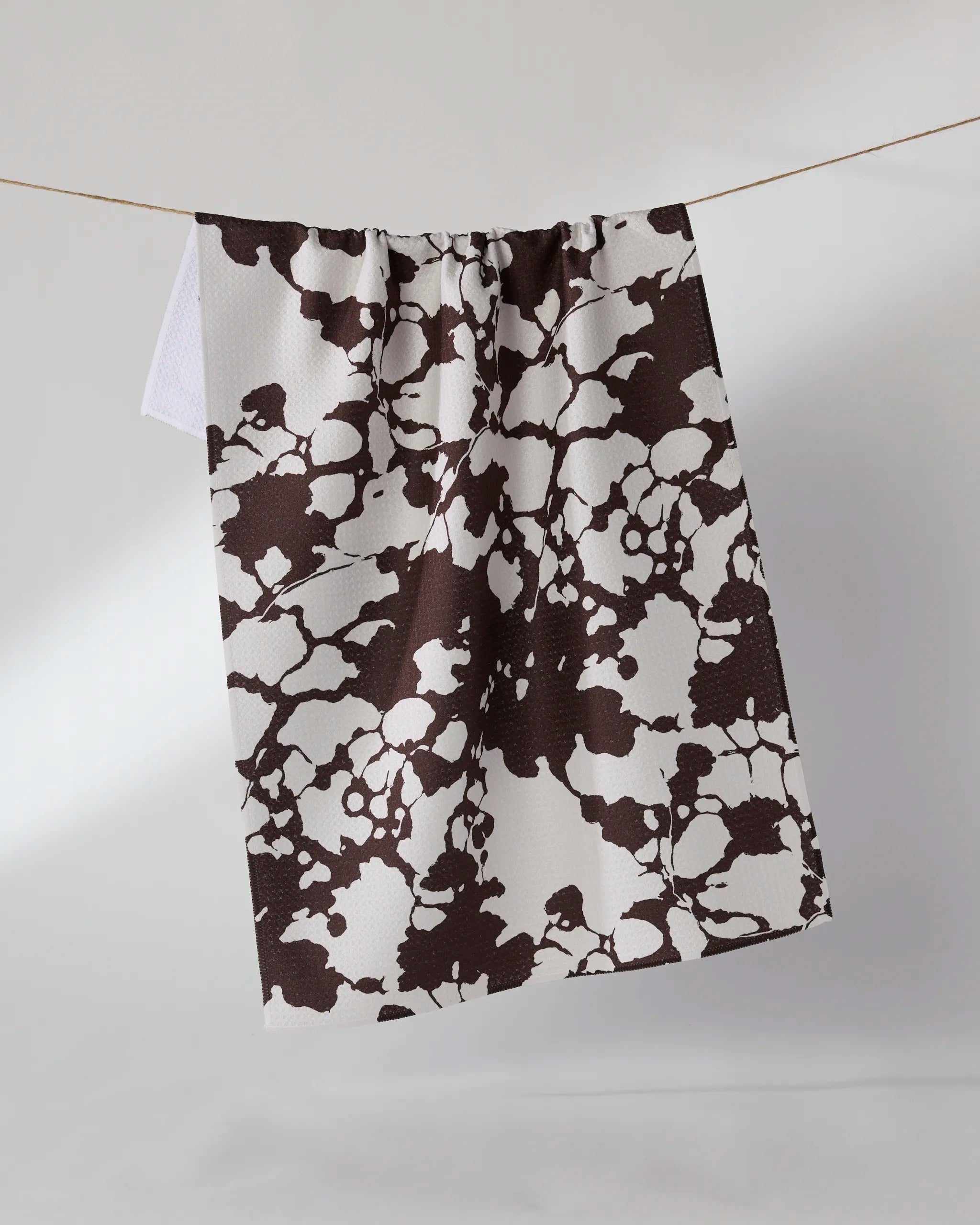 The Geometry Vine Noir towel, featuring an ultra-absorbent design and an abstract brown splatter pattern, hangs on a thin line against a plain light background.
