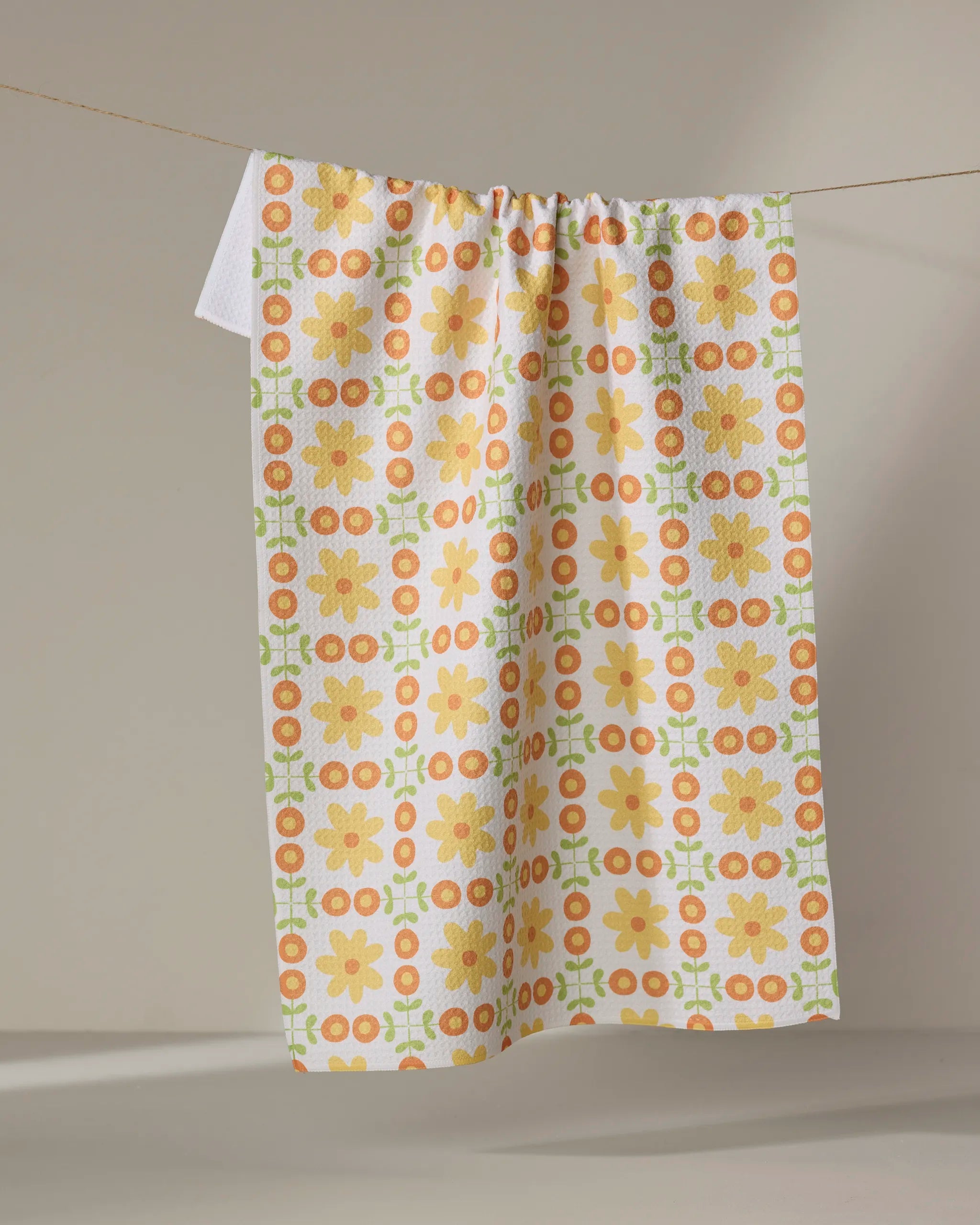 The Mindfulnice "Vintage Blooms" tea towel features orange and yellow flowers with green geometric shapes on white, hanging against a neutral backdrop. Made from ultra-absorbent recycled materials for eco-friendly use.