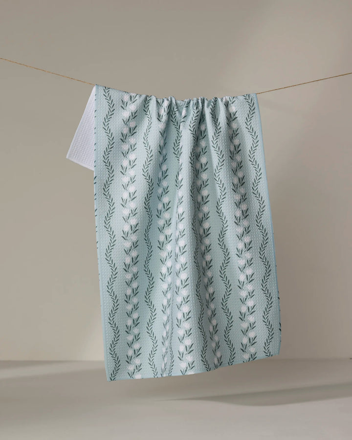 The Teri the Butter "Vintage Floral Stripe" tea towel, in light blue with a vertical leafy vine pattern, dries quickly as it hangs on a clothesline against a neutral background.