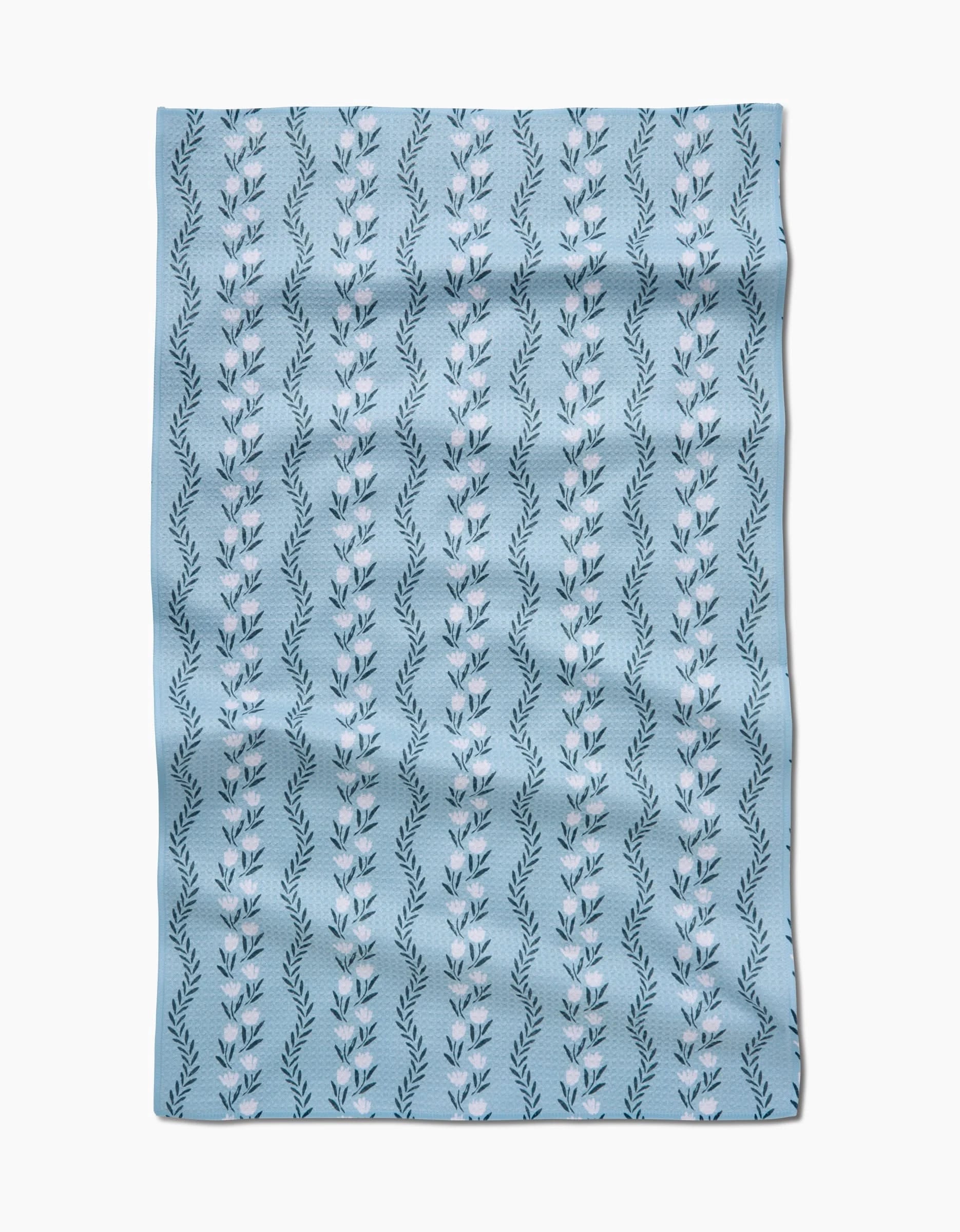 The Vintage Floral Stripe tea towel by Teri the Butter features light blue Geoweave fabric with vertical rows of white and pink stylized flowers and dark green leaves, shown slightly wrinkled against a white background.