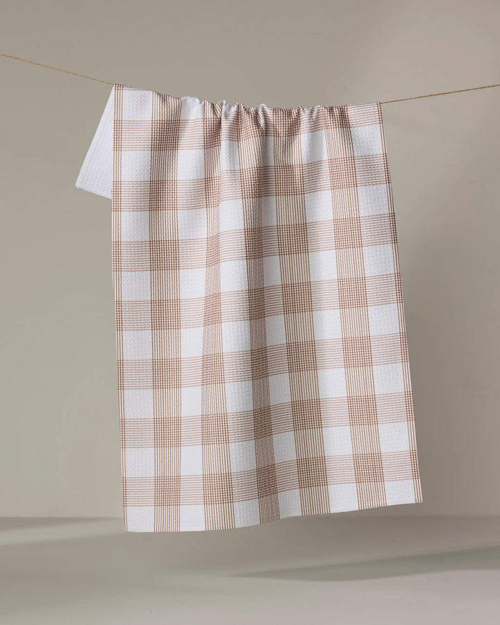 The Vintage Gingham Natural tea towel by Hufton Studio, crafted from recycled materials, features a light brown plaid on white and hangs on a string against a neutral background.