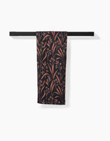 The Vintage Grass Black tea towel by Hufton Studio, featuring a brown and beige botanical pattern, hangs neatly folded over a modern black towel bar against a white background.