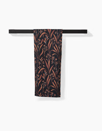 The Vintage Grass Black tea towel by Hufton Studio, featuring a brown and beige botanical pattern, hangs neatly folded over a modern black towel bar against a white background.