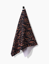 The Vintage Grass Black tea towel by Hufton Studio, featuring a brown and tan floral leaf pattern on dark recycled fabric, hangs from a black loop against a white background.