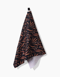 The Vintage Grass Black tea towel by Hufton Studio, featuring a brown and tan floral leaf pattern on dark recycled fabric, hangs from a black loop against a white background.