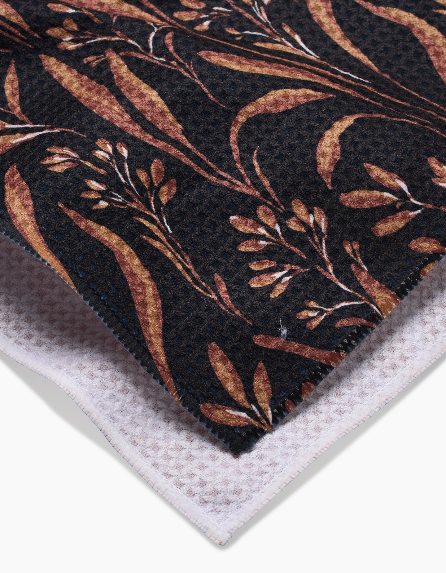 A close-up of two folded fabrics: the top is Hufton Studios Vintage Grass Black tea towel featuring a brown-beige botanical pattern, while the bottom is a textured white Geoweave waffle weave fabric.