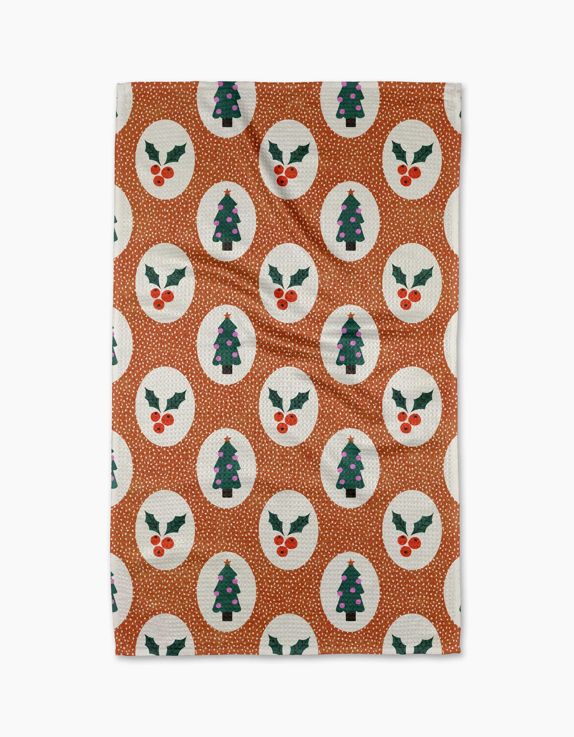 The Claire Iglesias Vintage Holly Christmas tea towel features a brown background with white circles displaying green Christmas trees with purple ornaments or holly leaves with red berries. Quick-drying, its both festive and practical.