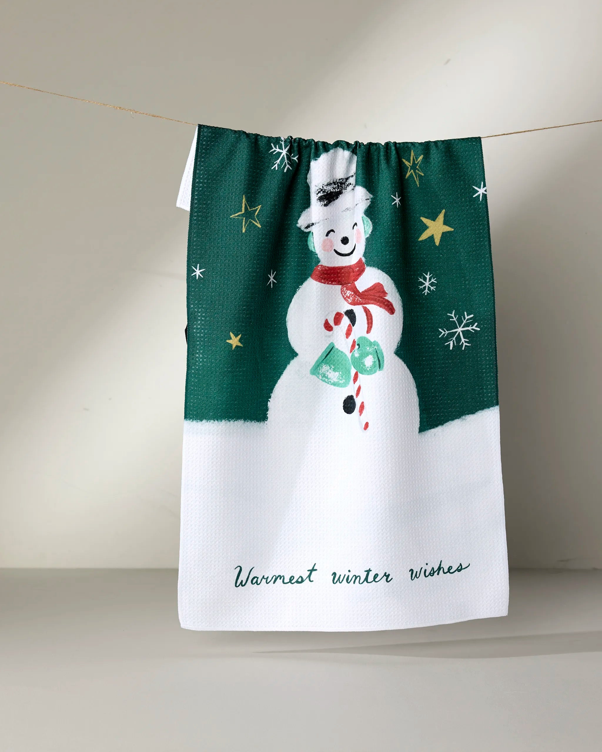 Add festive cheer to your kitchen with the "Vintage Snowman" towel by Louise Pretzel, featuring a smiling snowman with candy cane and wreath on green, gold stars and snowflakes, recycled materials, and "Warmest winter wishes" at the bottom.