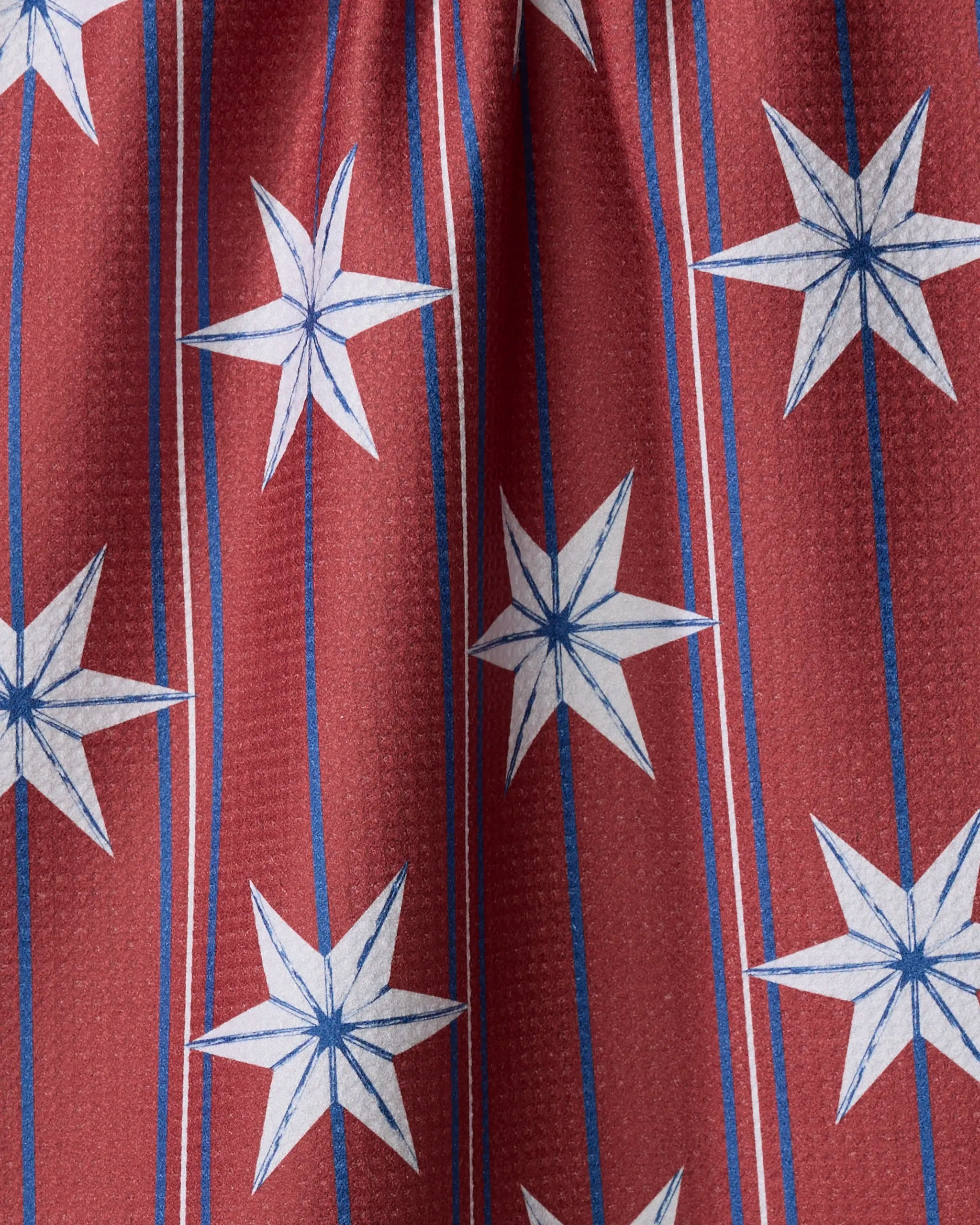 A close-up of Vintage Stars by Ashley Celena: red fabric with blue and white six-pointed stars and vertical stripes, slightly textured and gathered at the top—perfect as a festive, quick-drying tea towel for any kitchen.
