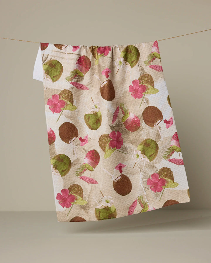 The Geometry "Vintage Tropi Drinks" kitchen towel, featuring a colorful pattern of coconuts, pink hibiscus, and palm leaves, hangs on a line against a neutral beige background.