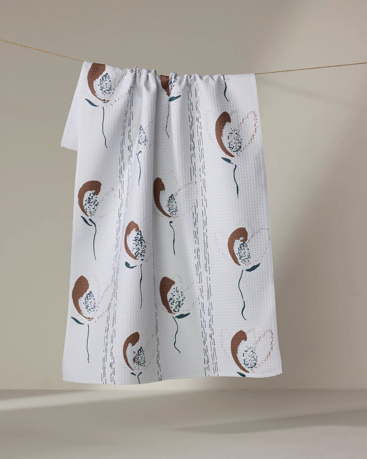 The "Vintage" kitchen towel by Toboggan Avenue features a white textured recycled fabric with brown, black, and blue floral prints, hanging on a clothesline against a neutral beige background.