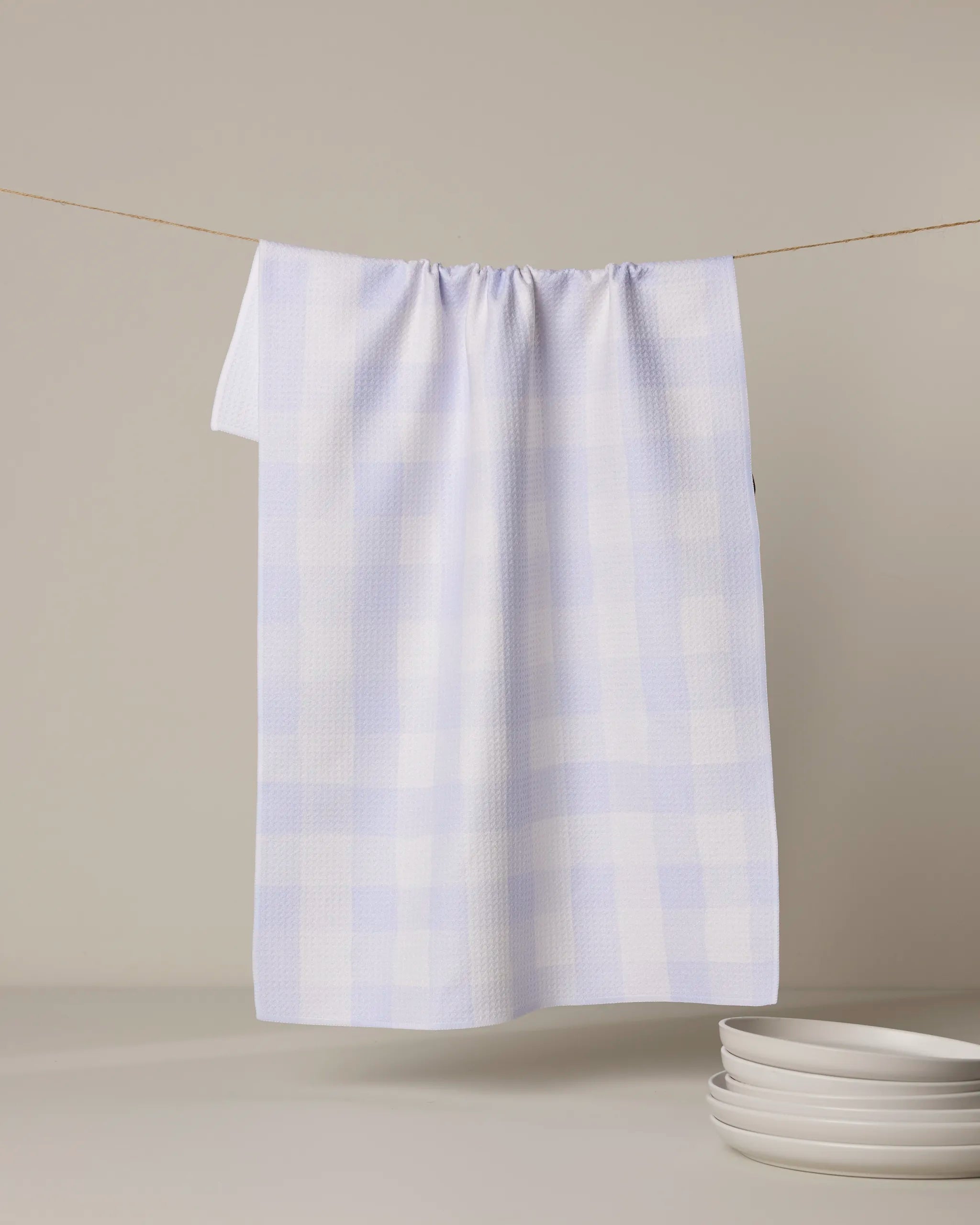 The Geometry Violetta tea towel, made from recycled materials in a light blue and white check pattern, hangs on a clothesline above stacked white plates against a neutral background.