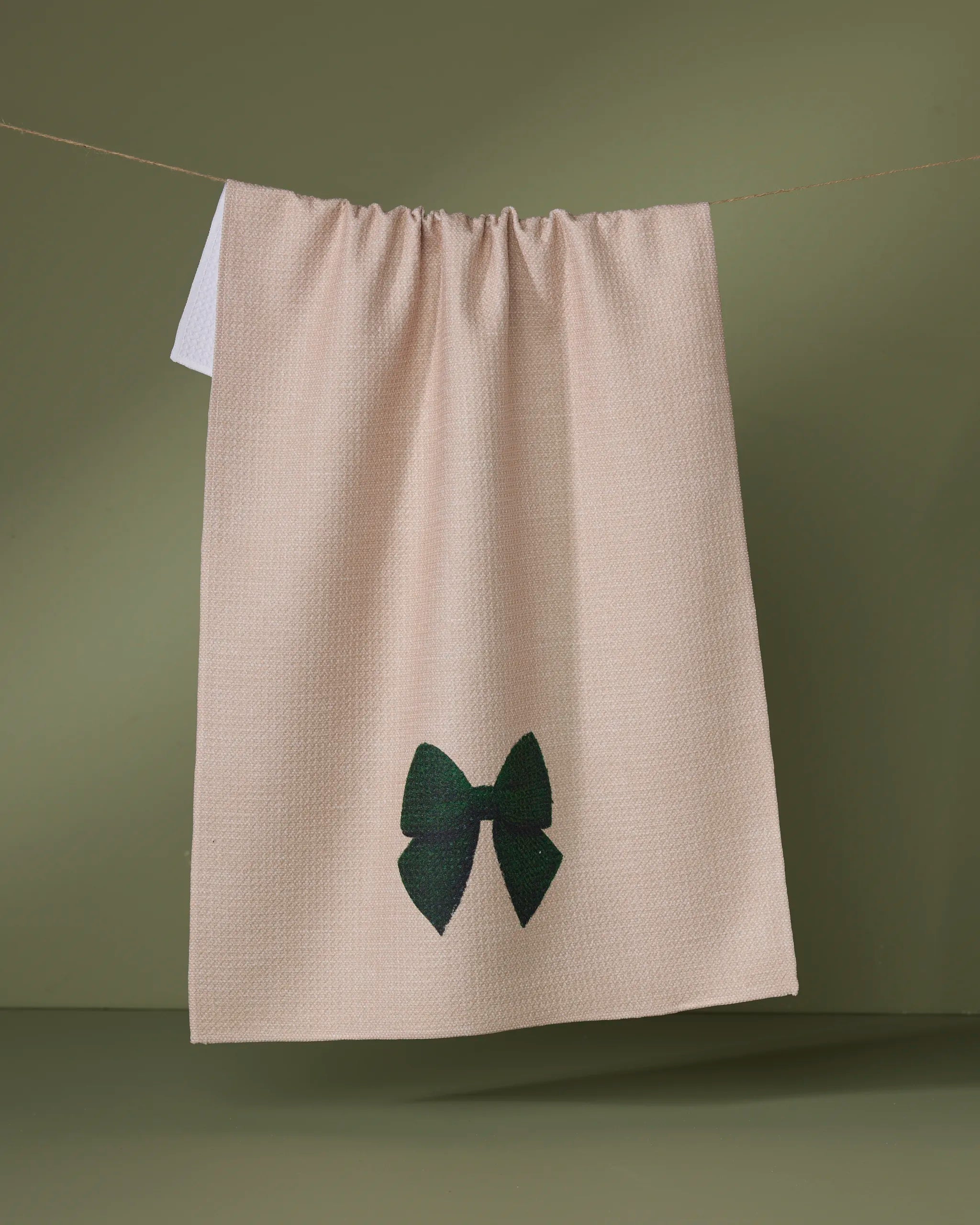 The Geometry Vireo dish towel, featuring a beige color and dark green embroidered bow, hangs on a clothesline with an olive green backdrop.