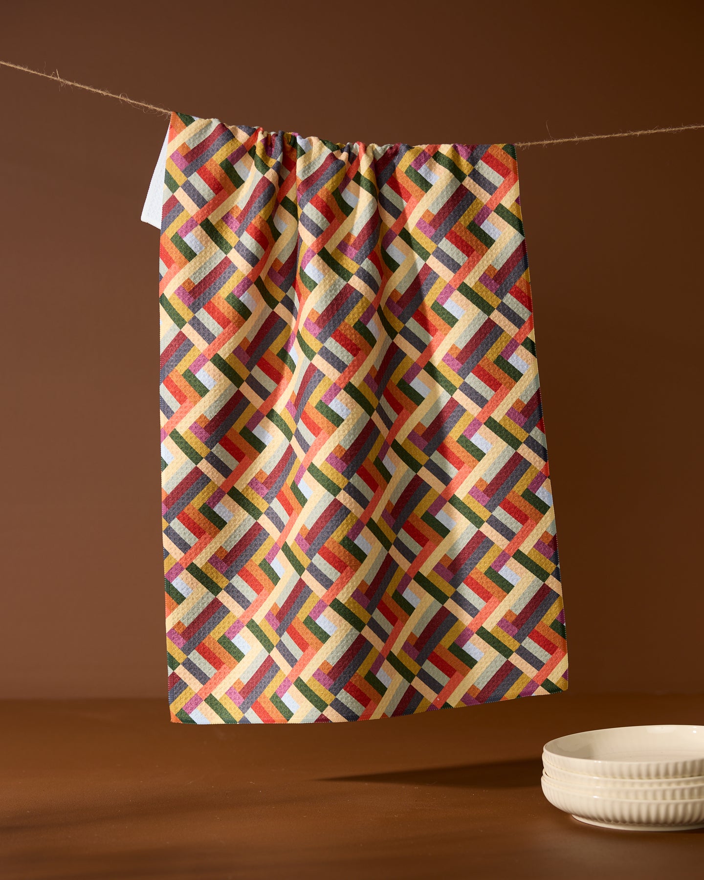 The Vivid Cascade tea towel by Geometry, featuring a vibrant geometric pattern, hangs from a string against a brown background with a stack of white plates in the lower right corner.