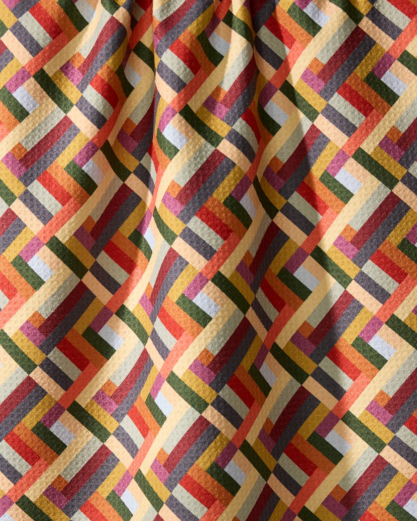 The Geometry Vivid Cascade kitchen towel features a vibrant, interlocking geometric pattern in yellow, red, green, orange, and blue. Made from recycled materials, its slightly wrinkled fabric creates gentle folds and subtle shadows.