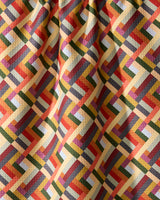 The Geometry Vivid Cascade kitchen towel features a vibrant, interlocking geometric pattern in yellow, red, green, orange, and blue. Made from recycled materials, its slightly wrinkled fabric creates gentle folds and subtle shadows.