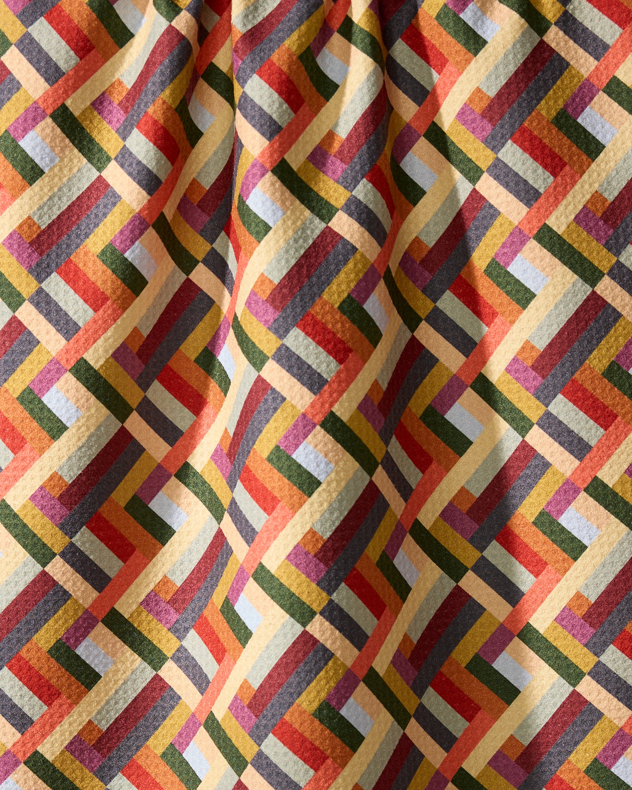 The Geometry Vivid Cascade kitchen towel features a vibrant, interlocking geometric pattern in yellow, red, green, orange, and blue. Made from recycled materials, its slightly wrinkled fabric creates gentle folds and subtle shadows.