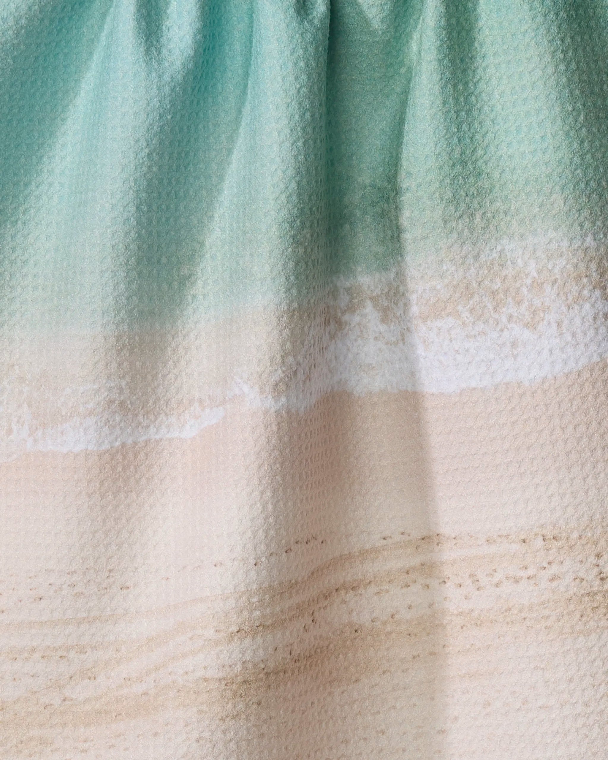 Close-up of the Waikiki kitchen towel by Kait Hanson, made from recycled fabric and printed with a beach scene featuring turquoise water, white waves, and beige sand with textured patterns. Fabric appears slightly wrinkled.