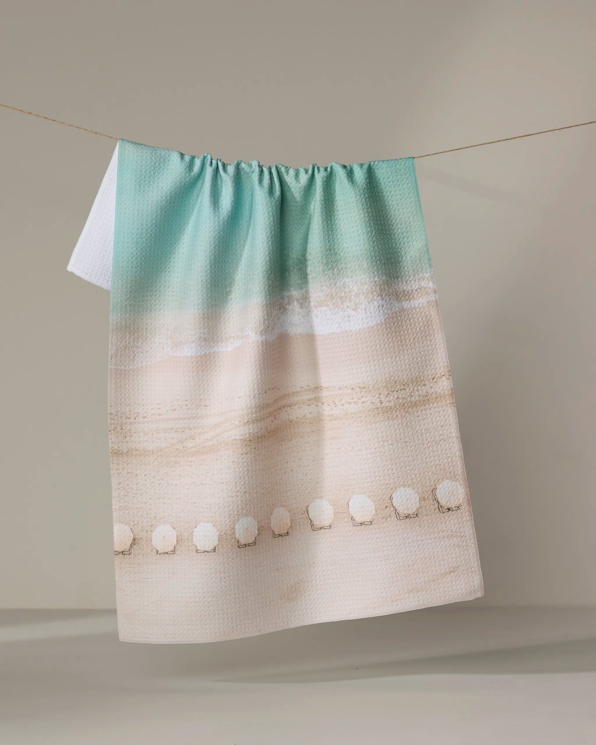 A quick-drying Waikiki towel by Kait Hanson hangs on a clothesline. It displays turquoise water, waves on sand, and white seashells along the bottom edge, set against a plain, neutral wall.