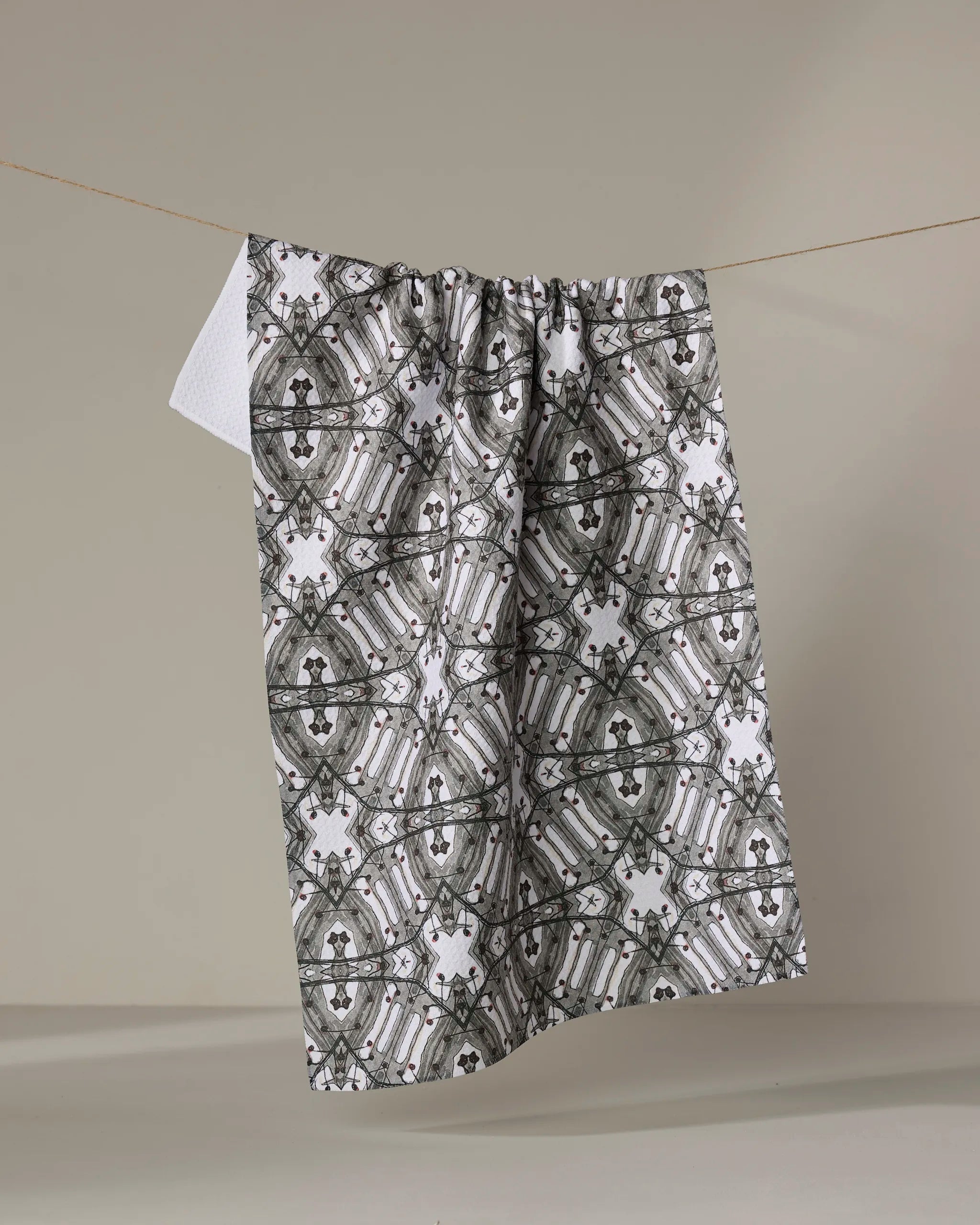 The Katherine Berkeley Walk The Line Shimmy towel, featuring an intricate black and gray symmetrical pattern, hangs on a clothesline against a plain light background with one corner revealing its white textured underside.