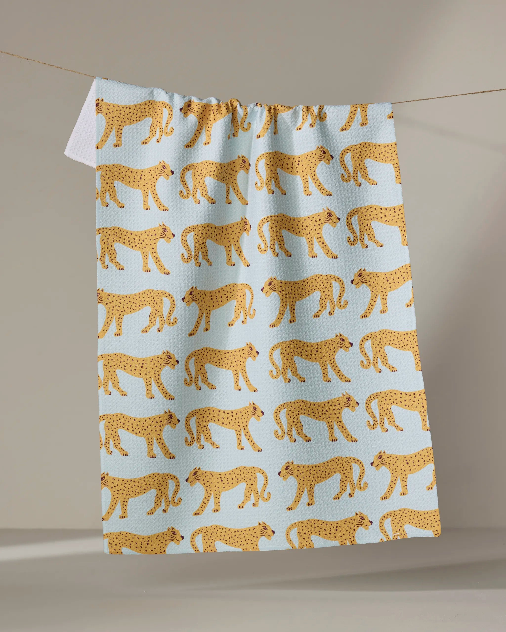 The Walking Cheetah tea towel by Jasmine Blooms Designs, in light blue with a repeating yellow cheetah pattern, hangs from a clothesline against a neutral background.