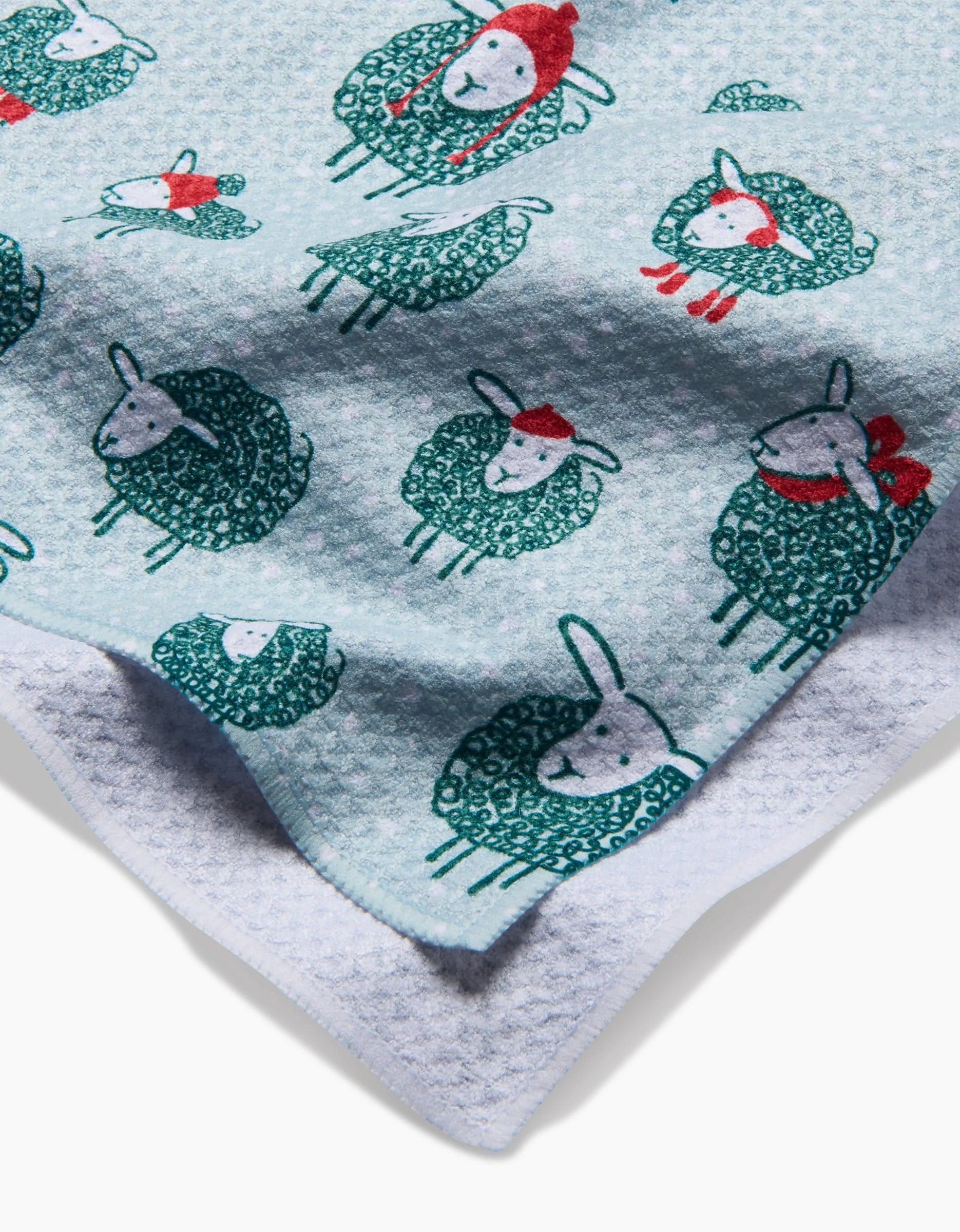 The Warm Woollysheep recycled kitchen towel by Kris Ruff features light blue textured fabric with playful sheep in red and green hats and scarves, plus a soft white fleece backing peeking at the corner.
