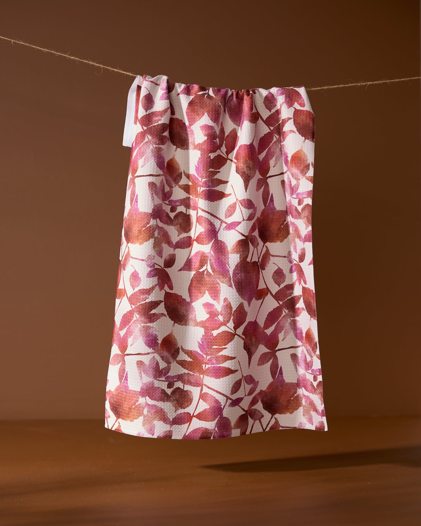 Geometry’s Watercolor Dreaming tea towel, featuring a pink and brown leaf pattern and made from recycled materials, hangs on a string against a brown background.