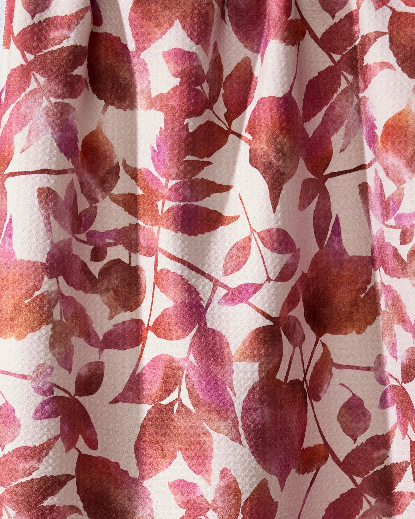 A close-up of Geometry’s Watercolor Dreaming tea towel, made from recycled materials, shows its pink, red, and brown leaf and flower pattern on a light background with a charmingly textured fabric.