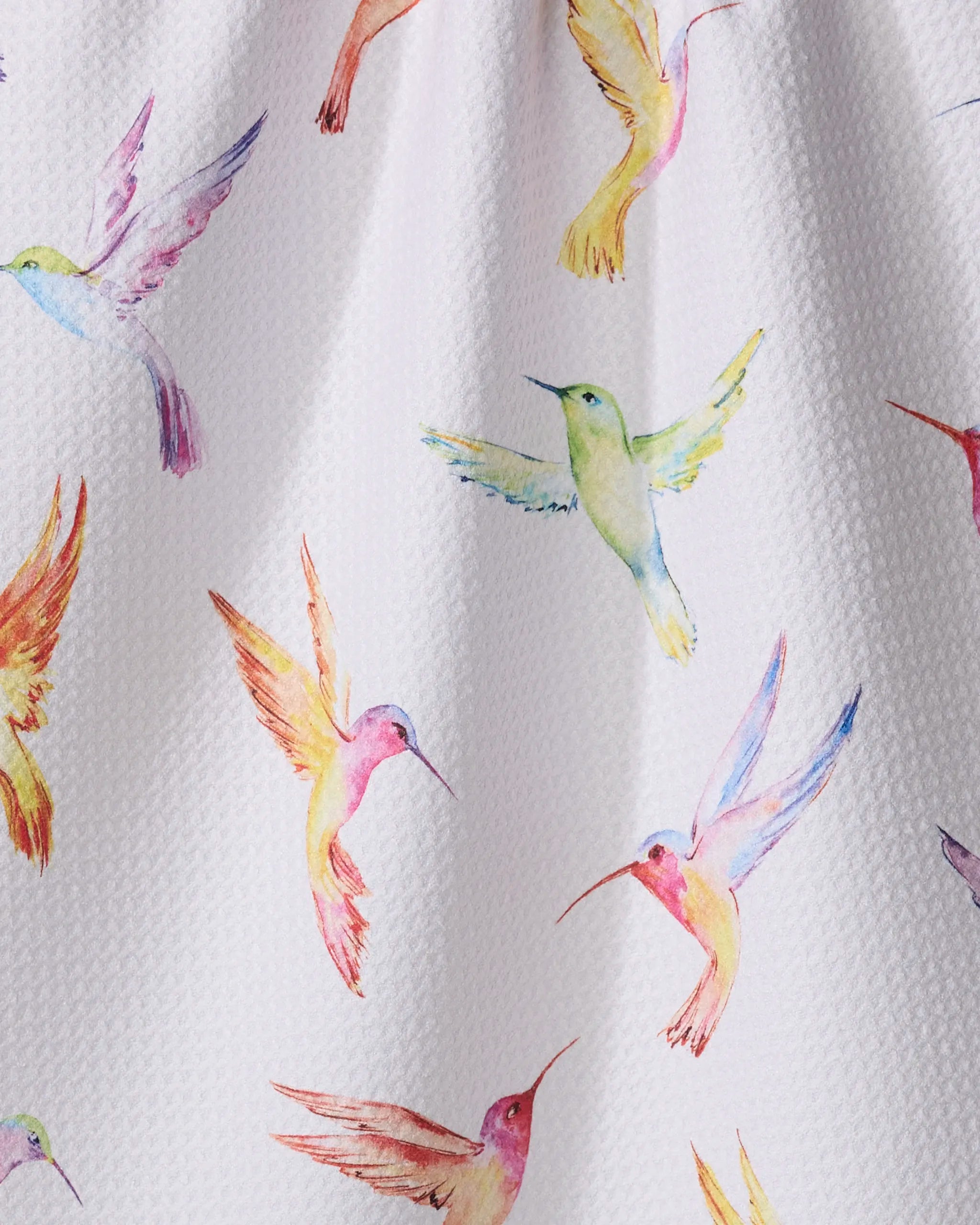 The Elena Fay Watercolor Hummingbirds kitchen towel features a lively pattern of pink, yellow, green, orange, and blue hummingbirds in flight on white textured fabric—perfect for brightening up your space.