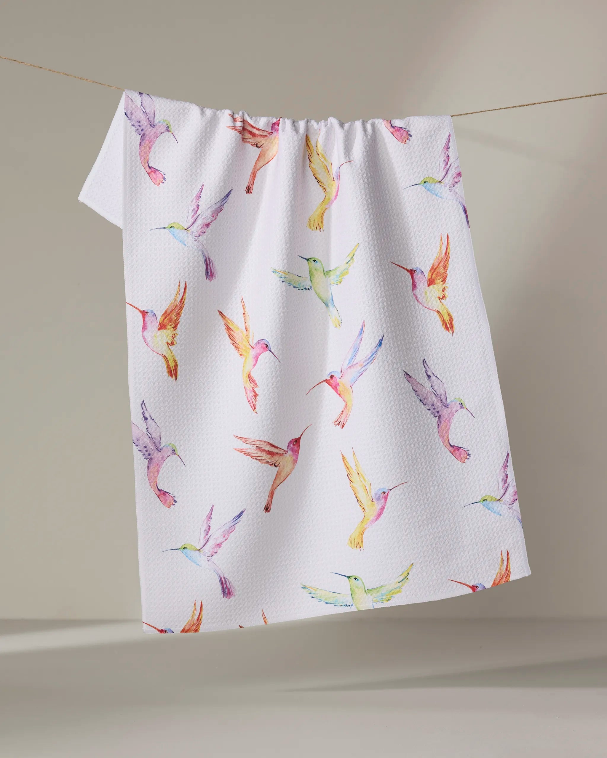 The Elena Fay "Watercolor Hummingbirds" kitchen towel, made from recycled materials and featuring a vibrant hummingbird design, hangs on a clothesline against a simple light background.