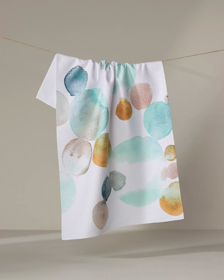 The Taryn Brennan Watercolor Rocks tea towel, made from recycled materials and featuring large teal, gold, peach, and beige watercolor-style circles, hangs on a line against a neutral background.